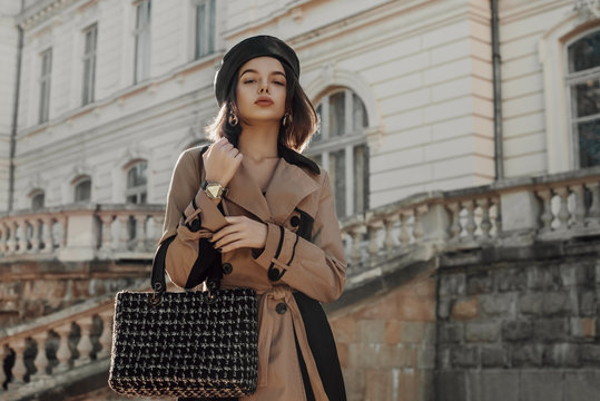 Outdoor autumn fashion portrait of young elegant woman wearing classic beige trench coat, trendy faux leather beret, holding black tweed bag, handbag, posing in street. Copy, empty space for text