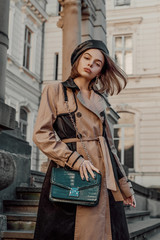 Outdoor autumn fashion portrait of young elegant confident woman wearing beige black trench coat, leather beret, holding green faux leather textured crocodile bag, posing in street of European city