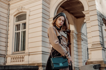 Outdoor autumn fashion portrait of young elegant woman wearing beige black trench coat, beret, with green faux leather textured crocodile shoulder bag, posing in street of European city. Copy space