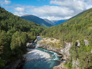 Naklejka na ściany i meble View on azure Rauma river canyon at Romsdalen valley with snow capped peaks of mountains, rocks and green forest. Blue sky white clouds background. Norway summer landscape scenery
