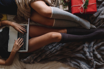 Two friends with knee high socks in winter holidays at home writing something on laptop near christmas tree in cozy interior with christmas presents.