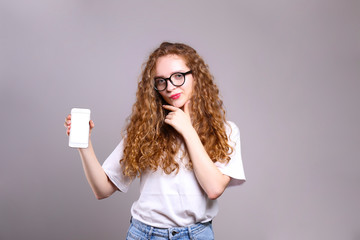 Young beautiful woman with long curly hair posing over isolated grey background & holding smartphone. Portrait of teenage female model wearing white t-shirt, showing emotions. Close up, copy space.