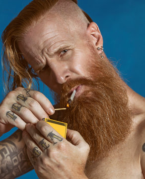 Thoughtful red haired bearded shirtless having tattoo on fingers and arms and with earrings in ear man with gray eyes and undercut hairstyle holding matchbox and lighting cigarette while frowning and looking at camera on isolated blue background