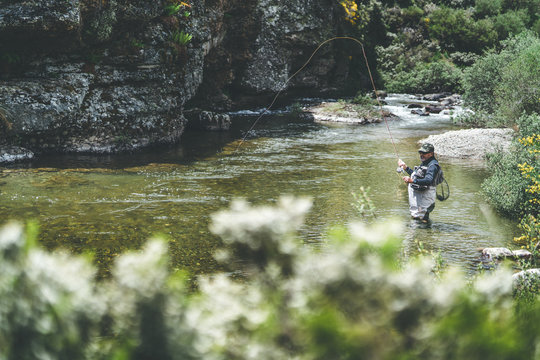 Side view of equipped man harling fish while standing in waders in mountain torrent by cliff and forest