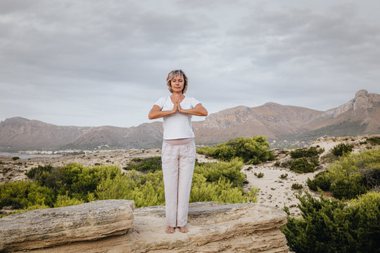 Full body barefoot adult female clasping hands over head and closing eyes while standing on rock in nature and meditating