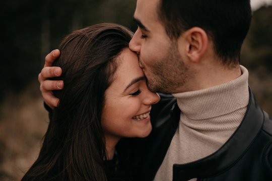 Side view of couple in love with closed eyes smiling while embracing and kissing each other along coniferous trees during daytime in windy overcast weather