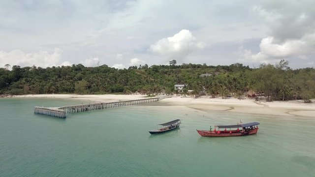 aerial panning shot of white sandy beach with long tail boats near wooden pier. Tropical rain forest as backdrop
