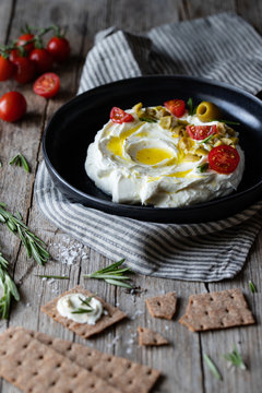 Crunchy crackers and rosemary placed on wooden table near napkin and plate of labneh yogurt with tomatoes and olives