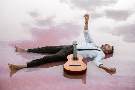 From above man in white shirt and suspenders lying on back looking away and floating with acoustic guitar and outstretching hand to the sky in sea at sandbank