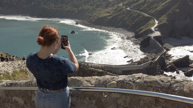 Young redhead girl taking a photo with a cellphone at San Juan de Gaztelugatxe, Spain. 4k and 50 fps