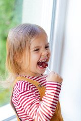 Portrait of cute toddler girl is sitting on windowstill with loly pop.