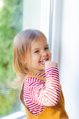 Portrait of cute toddler girl is sitting on windowstill with loly pop.