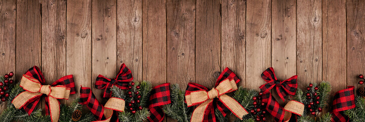 Christmas border banner with red and black checked buffalo plaid ribbon, burlap and tree branches. Above view on a rustic wood background.