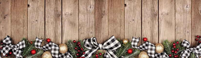 Christmas border banner with white and black checked buffalo plaid ribbon, ornaments and tree branches. Above view on a rustic wood background.