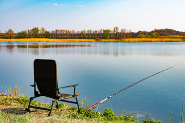 An empty chair and fishing rod on the lake coast in the morning. Green grass near large lake, blue sky, early hours