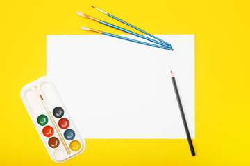 Blank paper with pencil, brushes and paints on yellow background. Artist workplace concept with copy space for your ideas. Flat lay, top view.