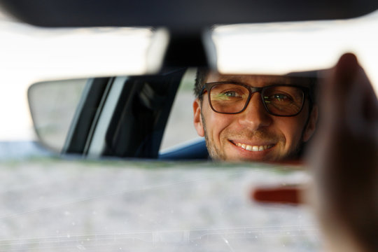 Cheerful joyfull man wearing glasses and adjusting mirror while sitting in his car, looking in reflection. soft focus. Emotions from driving a new car
