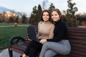 Beautiful two young twin sisters in casual outfit use notebook or laptop on bench at the park. Looking camera.
