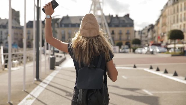 pretty girl, young woman with backpack and smartphone walks and dance along the alley with the flags and beautiful sky. Slow motion