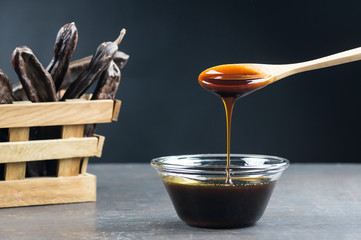 carob molasses in glass bowl and in wooden spoon and carob pods on rustic background, locust bean healthy food, Ceratonia siliqua ( harnup )