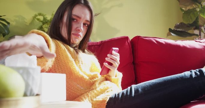 Unwell sick pretty caucasian young woman lying on couch with smartphone drinking hot tea warming up staying at home. Illness. Bad condition.