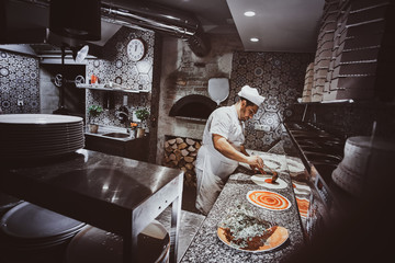 Italian chef in uniform is adding tomato sauce for pizza at the kitchen. - obrazy, fototapety, plakaty