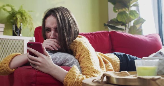 Young sick woman lying on sofa with smartphone sneezing allergy spitting mobile screen with snot showing disgust grimace. Illness concept.
