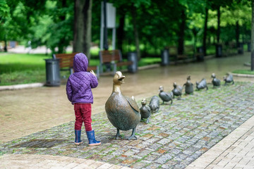 Fototapeta premium Unrecognizable little girl with her back to us counts the number of little ducklings of mothers duck, sculpture in the park