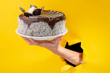 Hand holding a chocolate cake through a torn hole in yellow paper backgroun