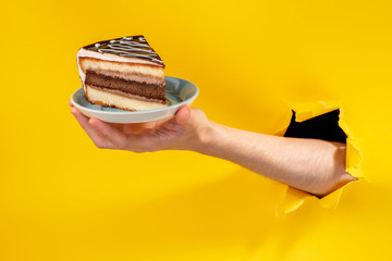 Hand offering a piece of cake through a torn hole in yellow paper background.