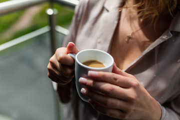 White mug with hot coffee inside the in the hands of women in Bathrobe standing on the balcony of her bedroom. Woman wearing white bathrobe with cup of coffee in her hands. Close up