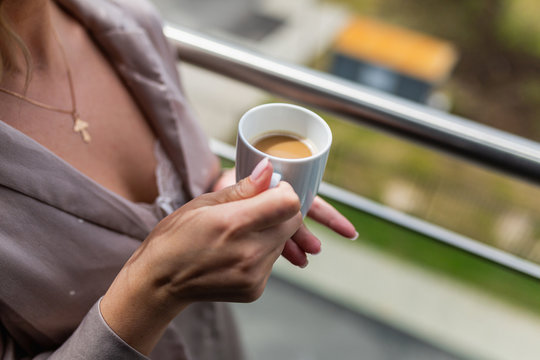 White mug with hot coffee inside the in the hands of women in Bathrobe standing on the balcony of her bedroom. Woman wearing white bathrobe with cup of coffee in her hands. Close up