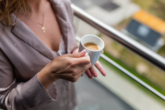 White mug with hot coffee inside the in the hands of women in Bathrobe standing on the balcony of her bedroom. Woman wearing white bathrobe with cup of coffee in her hands. Close up