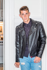 Portrait of Young Handsome Man at home in New York City. 20 years old man wearing black shirt, black leather jacket, blue ripped Denim jeans with broken holes, standing by white wooden door, looking..