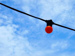 A round orange bulb close up hanging on a black wire On the blue sky background with white clouds...