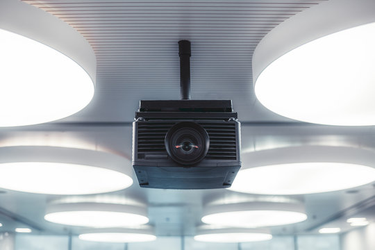 A Black Modern Projector With A Plastic Case And A Huge Lens With Aberrations In It Mounted To A Striped Ceiling With Multiple Huge Circle Ceiling Lamps Lighting A Modern Conference Hall