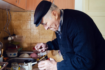 Senior man grandfather old pensioner farmer wearing black sweater and hat making a cup of coffee or tea cooking in the pot at home putting coffee to the pot with spoon