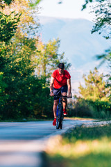 Obraz premium Fit cyclist rides his bicycle (bike) on an empty road in nature wearing a baseball hat and red t-shirt.