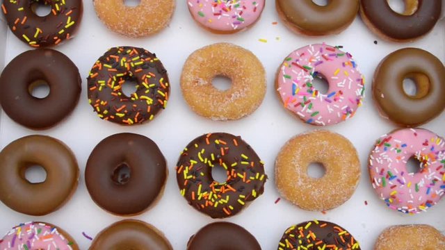 HD video background with 20 mini frosted donuts, various flavors and decorations rows in white bakers box. Young caucasian female hand reaches in and removes one donut as video zooms out to full box.