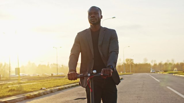 Young handsome black businessman riding electric scooter on empty country road at sunset. Close-up african man trying green transport. Ecology concept.