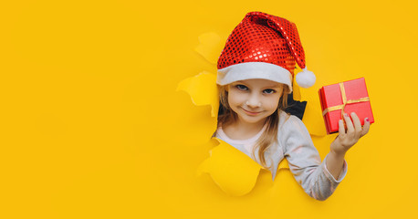 A cute little smiling girl in a red Santa hat looks out of a hole in yellow paper and holds a box with a gift in her hand. Christmas and New Year concept. Bright coloured background, copy space.