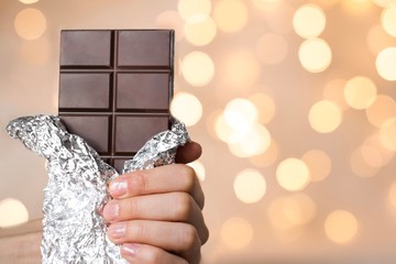 young woman holding a bar of a dark chocolate in a silver wrap with gold christmas lights on background