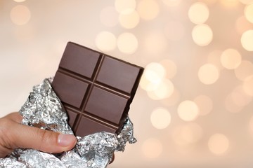 young woman holding a bar of a dark chocolate in a silver wrap with gold christmas lights on background