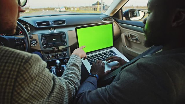 Multi-ethnic young business people driving in modern comfort car using mock-up laptop with greenscreen monitor running test drive program. Close-up. Luxury car interior.
