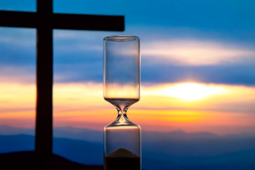 Hourglass has finished counting time on the background of the sunset and a wooden cross. End of...