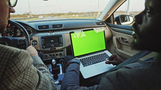 African man using modern laptop chrome key mock-up talking to colleague inside car. Couple of diverse friends driving car looking at green monitor laughing having fun.