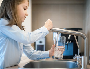 Portrait of a little caucasian girl gaining a glass of tap clean water. Kitchen faucet. Cute curly...