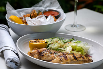 Grilled white fish with lemon and greens on a side and fried calamari rings with glass of white wine outside restaurant interior