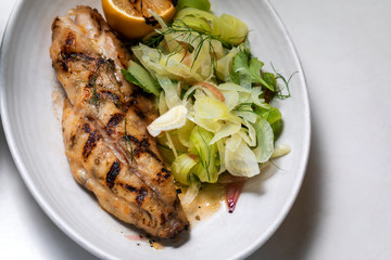 Grilled white fish with lemon and greens on a side outside restaurant interior
