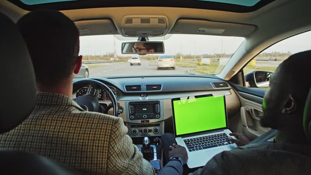 Two multi-race business colleagues driving in car to work using mock-up greenscreen laptop computer inside. Business people and technology.